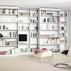 Eine moderne, weiße **eigene Bibliothek** mit vielen Büchern, Dekorationen und einem Fernseher. Ein Sessel und ein Hocker laden zum Verweilen ein.