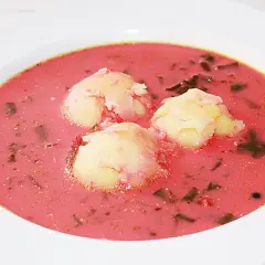 Pyszna zupa z botwiny z ziemniakami, podana w białej misce. Trzy kluski ziemniaczane unoszą się na różowym, aromatycznym bulionie.