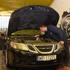 Mechanik sprawdza silnik czarnego Saaba. Na podnośniku warsztatowym, w tle widać narzędzia i części samochodowe.
