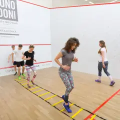 Grupa osób wykonuje dynamiczny stretching na drabince gimnastycznej w sali do squasha.