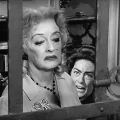 Bette Davis anziana, con un'espressione di disgusto, è dietro una grata. Un'altra donna con occhi sbarrati la spia.