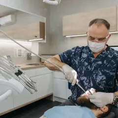 Dentysta w masce i okularach wykonuje zabieg, być może kortykotomię, u pacjenta leżącego na fotelu.