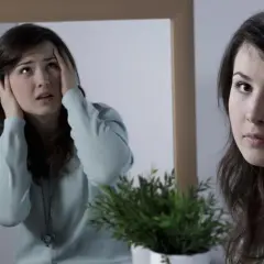 Młoda kobieta z niepokojem patrzy w lustro, gdzie jej odbicie chwyta się za głowę. Obraz symbolizuje walkę z objawami schizofrenii.