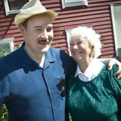 Sigge Dorsin ler mot kameran med en äldre kvinna. De står framför ett rött hus.