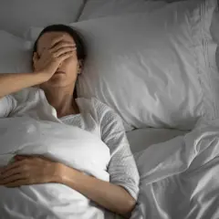 Frau liegt im Bett, bedeckt mit einer Decke, und hält sich die Hand vor die Augen. Sie kann nicht schlafen.