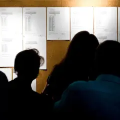 Sylwetki ludzi patrzą na tablicę ogłoszeń z listami wyników rekrutacji do szkół.