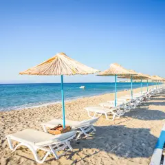 Słoneczna plaża na wyspie Kos, rzędy leżaków pod słomianymi parasolami, turkusowe morze i błękitne niebo. Idealne miejsce na relaks.
