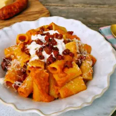Un plato de macarrones con chorizo, cubiertos con queso rallado y una cucharada de crema. Al fondo, tomates y un trozo de queso.