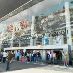 Ludzie z bagażami przed nowoczesnym budynkiem Aeroporto Internazionale di Napoli.
