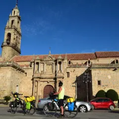 Ciclistas preparan sus bicicletas para continuar las etapas de Transandalus frente a una iglesia histórica bajo un cielo azul.