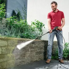 Mężczyzna w czerwonej koszulce i jeansach czyści kamienny murek myjką ciśnieniową.