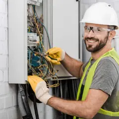 Uśmiechnięty technik elektryk w białym kasku i żółtych rękawicach pracuje przy skrzynce z przewodami.