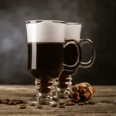 Två glas med kaffe med glass och vispgrädde, omgivna av kaffebönor och kanelstänger på ett träbord.