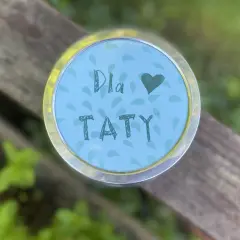 Uroczy upominek na Dzień Taty z przedszkola. Na okrągłej, błękitnej nakrętce z listkami, ręcznie napisane "Dla Taty" i serduszko.