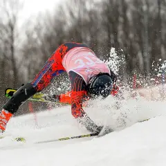 Narciarz w czerwonym kombinezonie traci równowagę, śnieg wzbija się w powietrze. Widoczny obciążenie tyłów nart przy skręcie.