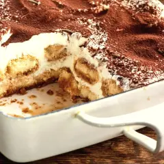 Tiramisu w białej formie, posypane kakao, z widocznymi warstwami biszkoptów i kremu.