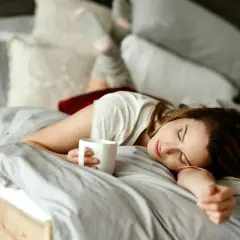 En kvinna sover i sängen med en kaffekopp i handen. Kaffe bra eller dåligt? Kanske en tupplur är bättre.