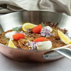 En gyllene råraka i stekpanna, toppad med gräddfil, rödlök, rom och citronklyfta. Ett klassiskt råraka recept som ser utsökt ut.