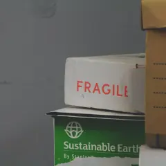 Paczka przyjęta w oddziale. Na białym kartonie napis "FRAGILE", pod nim zielony z napisem "Sustainable Earth". Obok brązowe pudło.