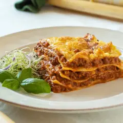 Porción de lasaña de carne casera, con capas de pasta, salsa boloñesa y queso gratinado, servida con brotes y albahaca.