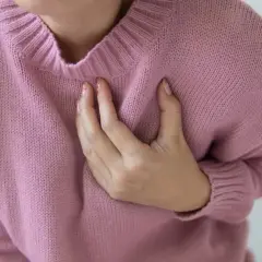 Kobieta w różowym swetrze odczuwa kłucie pod lewą piersią, trzymając się za serce.