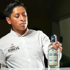 Młody mężczyzna w białej koszuli prezentuje butelkę tequili 30-30 Reserva Especial Blanco, podkreślając, z czego jest tequila – ze 100% niebieskiej agawy.