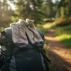 Ett par slitna men varma ullstrumpor vilar på en ryggsäck vid en solbelyst skogsstig. Perfekt för vandring med bra ull.