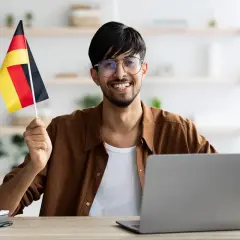 Uśmiechnięty mężczyzna z flagą Niemiec przy komputerze, planuje otwarcie konta w niemczech bez meldunku.
