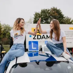 Dwie uśmiechnięte dziewczyny świętują sukces po egzaminie praktycznym kat B. Siedzą na dachu samochodu nauki jazdy, otoczone innymi pojazdami szkoleniowymi.