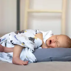 Śpiące niemowlę otulone kocykiem leży na szarej poduszce stabilizacyjnej.