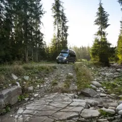 Ciemny kamper na leśnej drodze, otoczony przez wysokie drzewa i kamieniste koryto strumienia. Idealne miejsce na **split camping** i ucieczkę od cywilizacji.