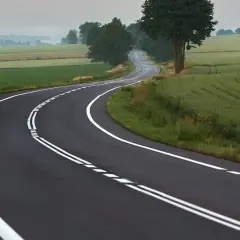 Zakręt drogi z podwójną ciągłą linią, gdzie wyprzedzanie jest zabronione. Pola i drzewa w tle.