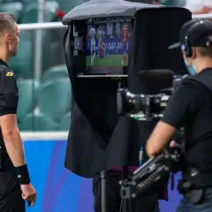 Sędzia obserwuje powtórkę na monitorze podczas meczu piłki nożnej. W tle operator kamery.