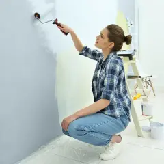 Młoda kobieta w kratę i jeansach maluje ścianę wałkiem. W tle drabina, puszki z farbą i krzesła.