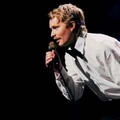 En man med mustasch, klädd i vit skjorta, sjunger i mikrofonen under nalles show.