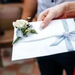 Dłoń trzyma białą kopertę z bukiecikiem i błękitną wstążką. Zastanawiasz się, ile chrzestny daje na wesele?