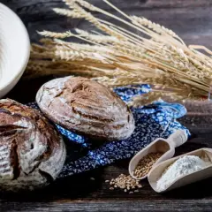 Zwei rustikale Brote, Weizenähren und Holzschaufeln mit Getreide und Mehl. Eine Bäckerei-Szene, die die Vielfalt einer **Getreide Sorte** zeigt.