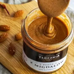 Gęsta, kremowa pasta z migdałów, czyli polskie "noisette", w słoiku, otoczona migdałami na drewnianej desce.