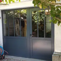 Remplacer porte de garage par fenêtre : une nouvelle entrée lumineuse avec des portes vitrées grises. Des outils de jardin sont rangés à gauche.