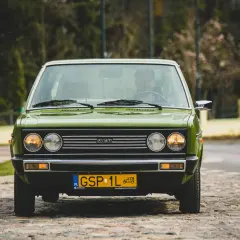 Zielony Fiat 131 Mirafiori z lat 70. na brukowanej ulicy, z widocznym kierowcą za kierownicą.