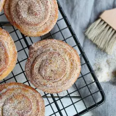 Nybakade kanelbullar med strössel av brunt socker och kanel på ett svart galler. En pensel och en sked med socker ligger bredvid.