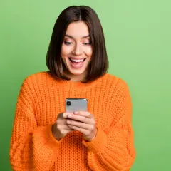 Uśmiechnięta kobieta w pomarańczowym swetrze trzyma smartfon. Może właśnie dostała informację o wyłączeniu 3G Orange i cieszy się z nowej oferty.