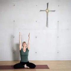Frau in Yoga-Pose mit erhobenen Armen vor einem Kreuz an einer Betonwand. Eine harmonische Verbindung von Yoga und Christentum.