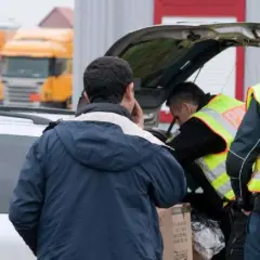 Polizisten durchsuchen ein Auto. Ein Mann telefoniert, während die Beamten den Kofferraum inspizieren. Im Hintergrund stehen Lastwagen.
