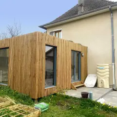 Une extension en bois moderne s'intègre à une maison existante, avec de grandes fenêtres et une finition en lattes de bois.