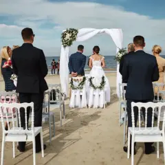 Piękny ślub na plaży. Para młoda stoi przed białym baldachimem, goście obserwują ceremonię.