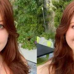 Mujer con cabello largo y ondulado en tonos tinte pelirrojo, ojos azules y pecas, sonríe en un entorno exterior con vegetación.