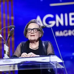 Agnieszka Holland w okularach za mównicą z mikrofonem i statuetką.