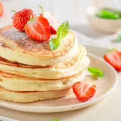 Puszyste ciasto na pancake, ozdobione świeżymi truskawkami i listkami mięty, wygląda apetycznie.