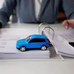 Osoba z długopisem i kalkulatorem analizuje dokumenty, obok stoi niebieski model auta. Zastanawia się, po jakim czasie można sprzedać auto.
